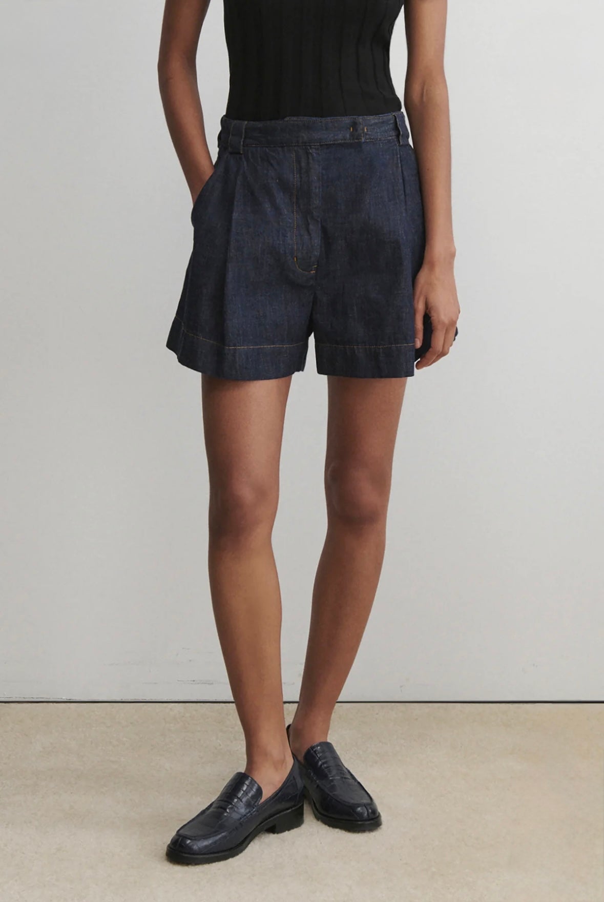 Person wearing dark denim shorts and black shoes on a plain background